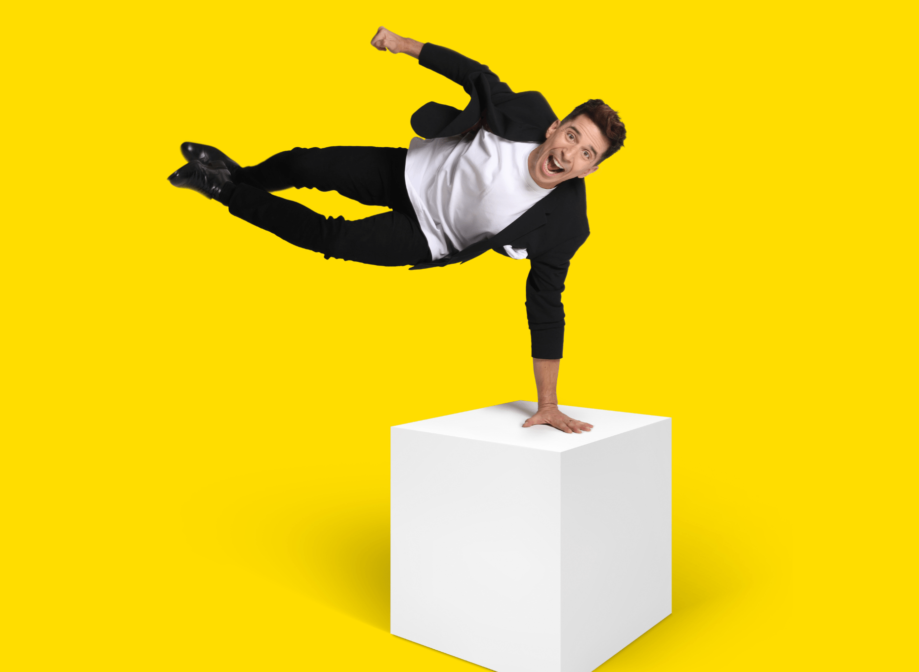 Comedian Russell Kane balances on a white cube with one hand holding his entire body in the air. He is wearing a black suit and a white t-shirt and has a joyful expression on his face.
