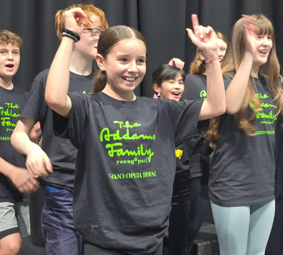A group of excited kids in matching "The Addams Family" T-shirts perform on stage, smiling and raising their hands joyfully, conveying enthusiasm.