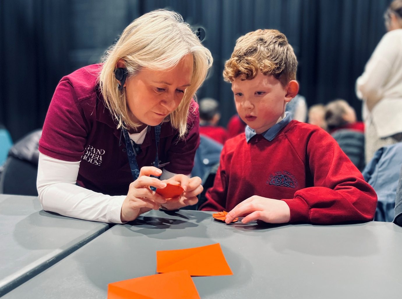 A woman in a red Grand Opera House t-shirt assists a young boy with a craft project using orange paper. They are focused and seated at a grey table with other children and adults working on crafts in the background.