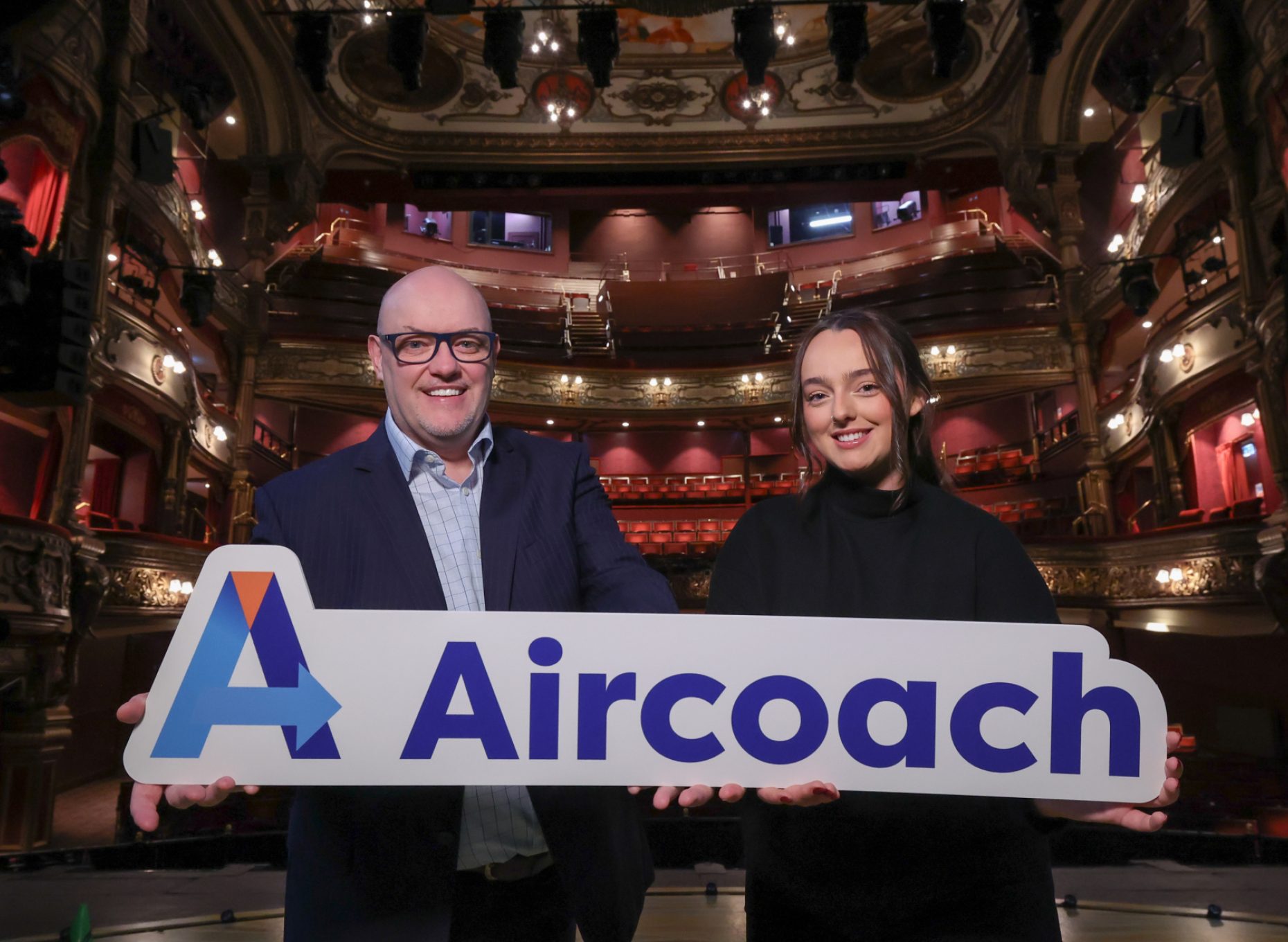 Ian Wilson Grand Opera House Chief Executive and Emily Dover, Aircoach Marketing Executive on the Grand Opera House stage  both holding one end of an Aircoach logo prop with the Auditorium in the background.