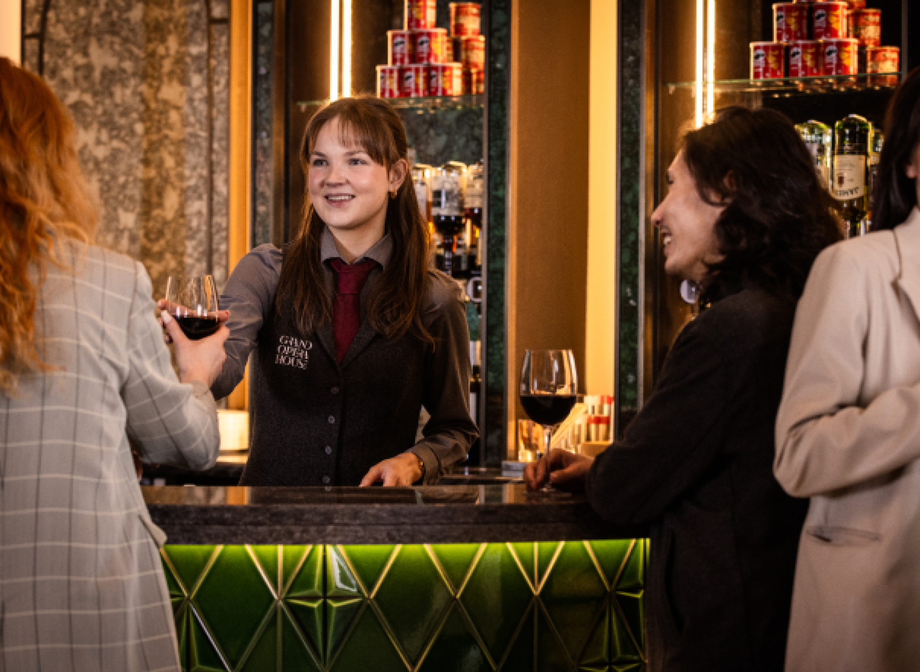 A server passes a glass of red wine to a woman across a car. The server is smiling and wearing a red tie and grey shirt/waistcoat. A man with long hair stands at the bar drinking a glass of wine and smiling.