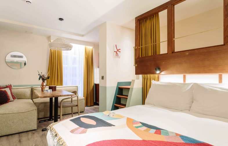 A cosy hotel room featuring a neatly made bed with a colourful patterned blanket, mustard-yellow curtains, a small dining area, and modern décor.