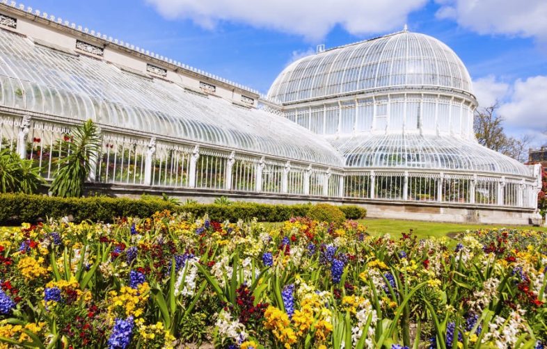 Belfast's botanical gardens can be seen, in the foreground are multiple colourful flowers and in the background is a large ornate greenhouse building. The sky is blue with some white clouds.