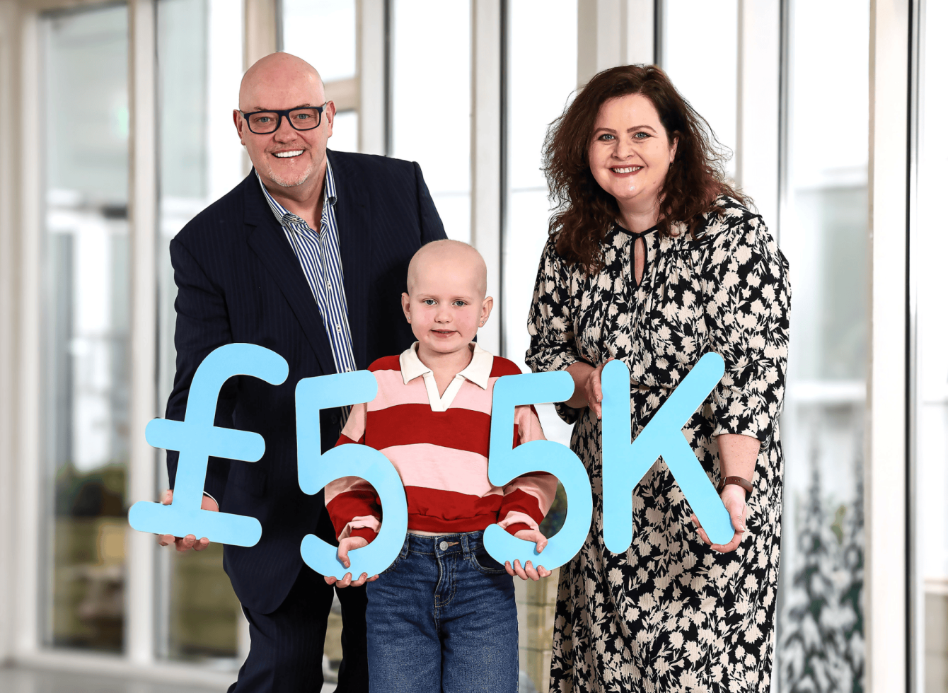 A child in a red striped shirt is flanked by a smiling man in a suit and a woman in a floral dress. They hold a large "£55K" sign indoors.