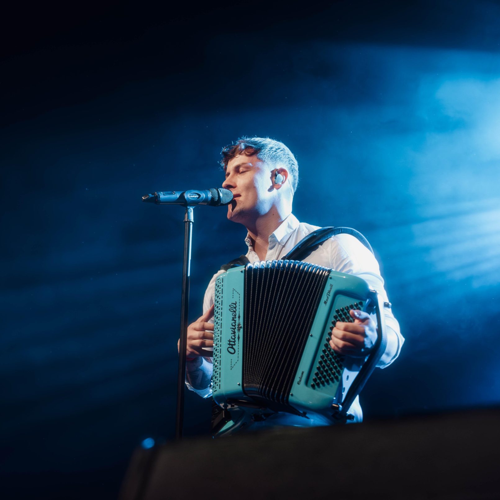 A man passionately sings and plays an accordion on stage under dramatic blue lighting, creating a serene and soulful atmosphere.