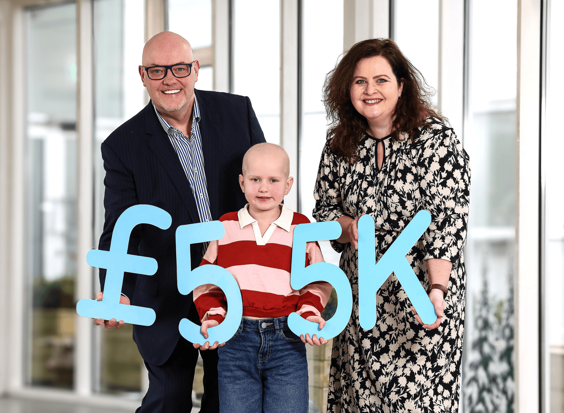 A child in a red striped shirt is flanked by a smiling man in a suit and a woman in a floral dress. They hold a large "£55K" sign indoors.