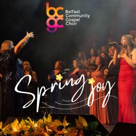 Banner reading “Belfast Community Gospel Choir” above a choir performing onstage with raised hands and yellow flowers in front.
