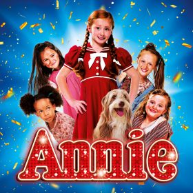 Young girl in a red dress with braided hair, surrounded by five children and a dog, smiles brightly under confetti on a blue background. Text reads "Annie."