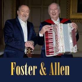 Two older men in dark suits; one holds a guitar, the other an accordion. They smile warmly in an elegant room. Text below reads "Foster & Allen."