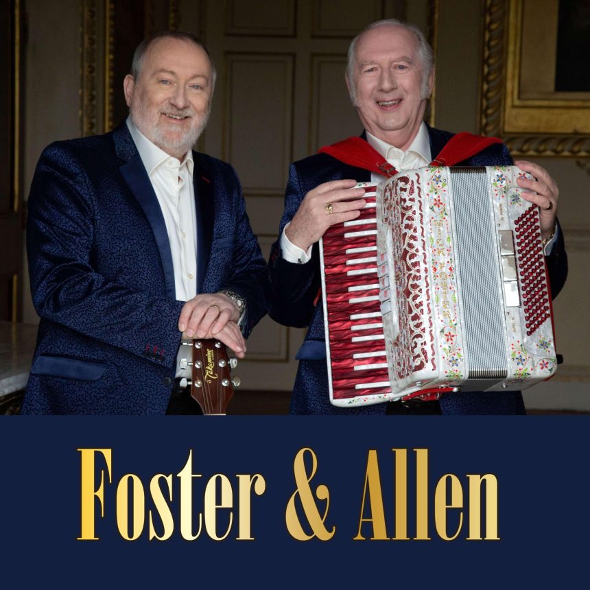 Two older men in dark suits; one holds a guitar, the other an accordion. They smile warmly in an elegant room. Text below reads "Foster & Allen."